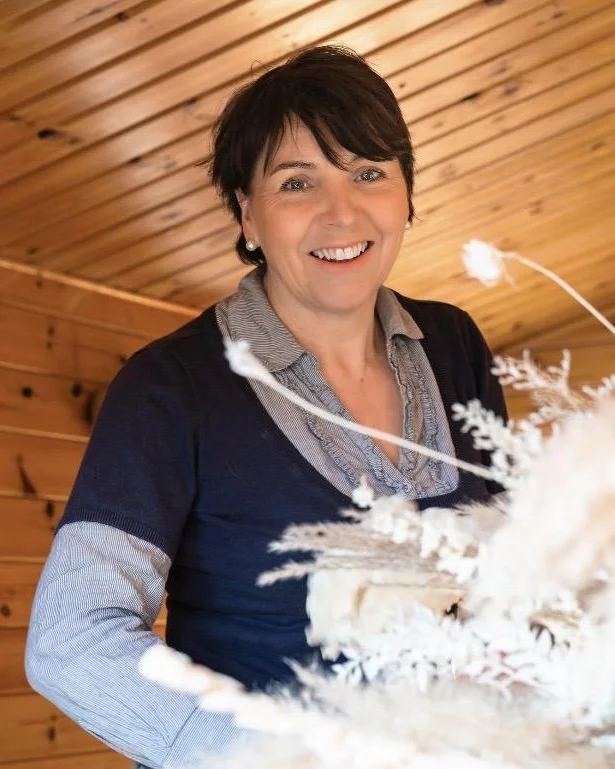 Catherine, a dried flower florist in Dorset, smiling behind a large arrangement of pale dried grasses.