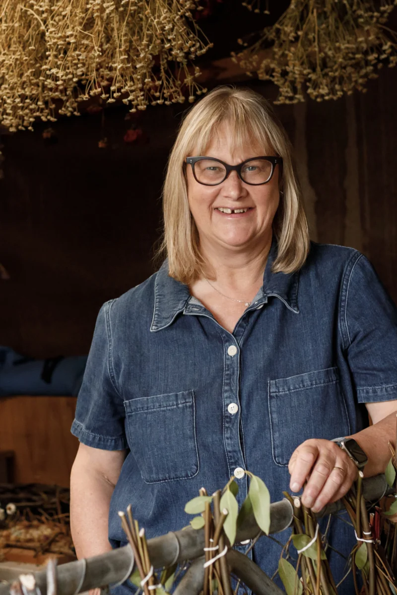Julie Treanor, a flower grower in New Zealand, smiling in a denim shirt beneath hanging dried flowers.