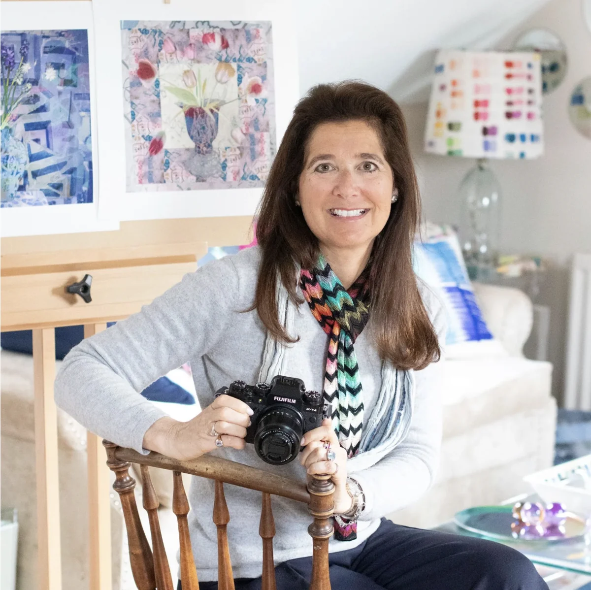 Juliette, a photographic artist, smiling in her studio with a camera, her paintings on the wall behind her.