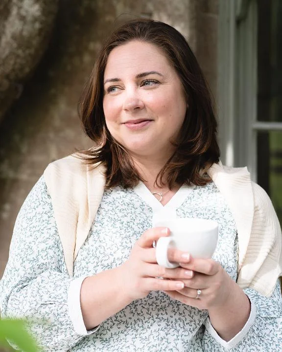 Keri, a garden designer, holding a cup of tea in a garden beside an old tree.