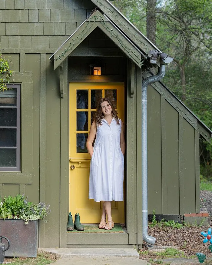Molly, a garden designer in Alabama, smiling in the yellow doorway of a green shingled cottage among trees.