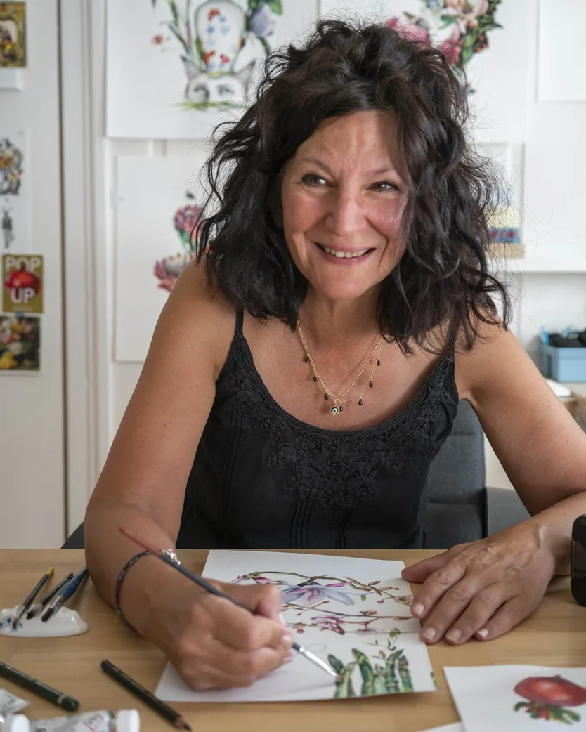 Tina Stamilo, an illustrator in Switzerland, smiling at her desk with a pencil in hand, painting botanical illustrations.